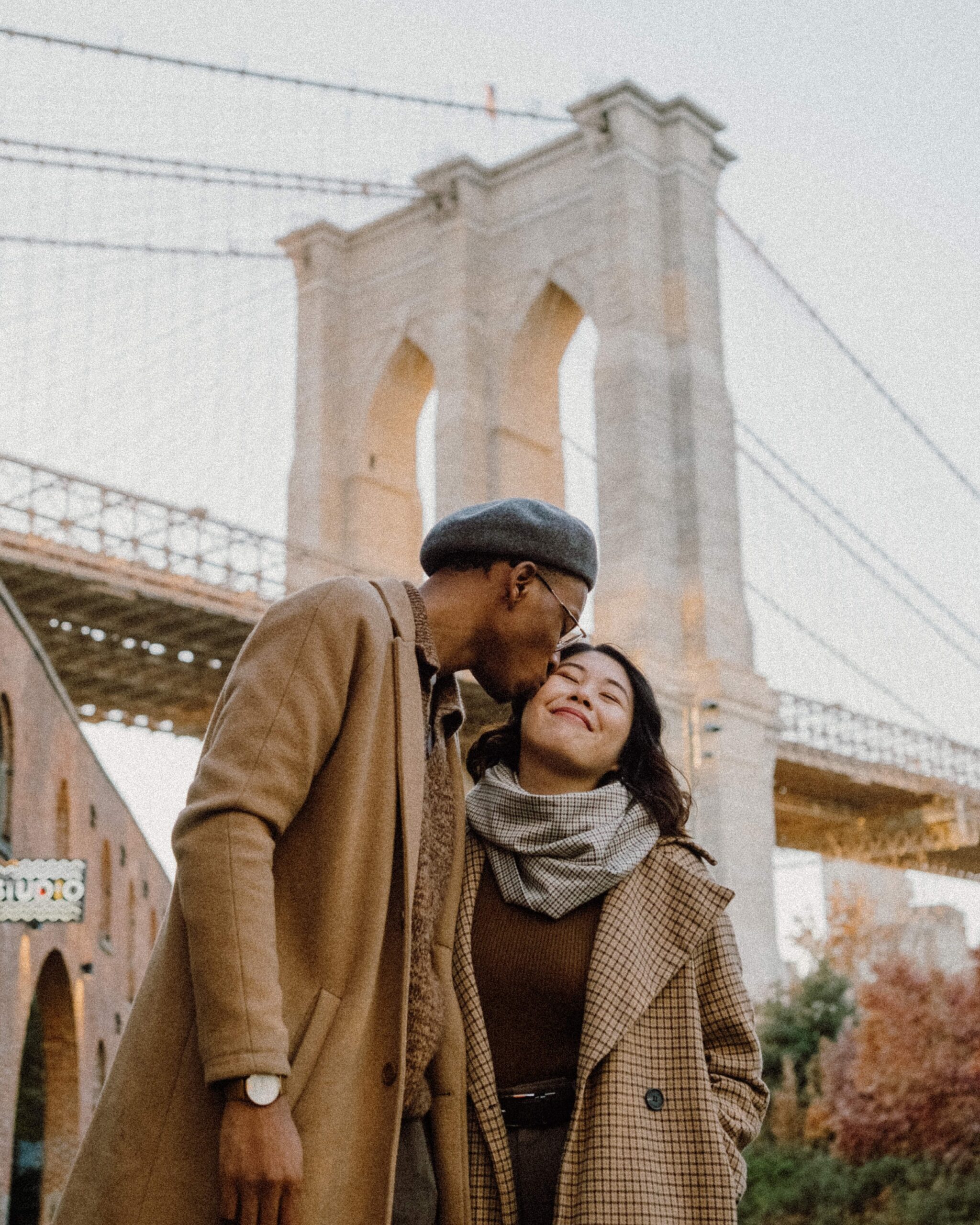 S & D standing together in Dumbo, New York City during their engagement session — soft golden light, cobblestone streets, and the Brooklyn Bridge in the background.